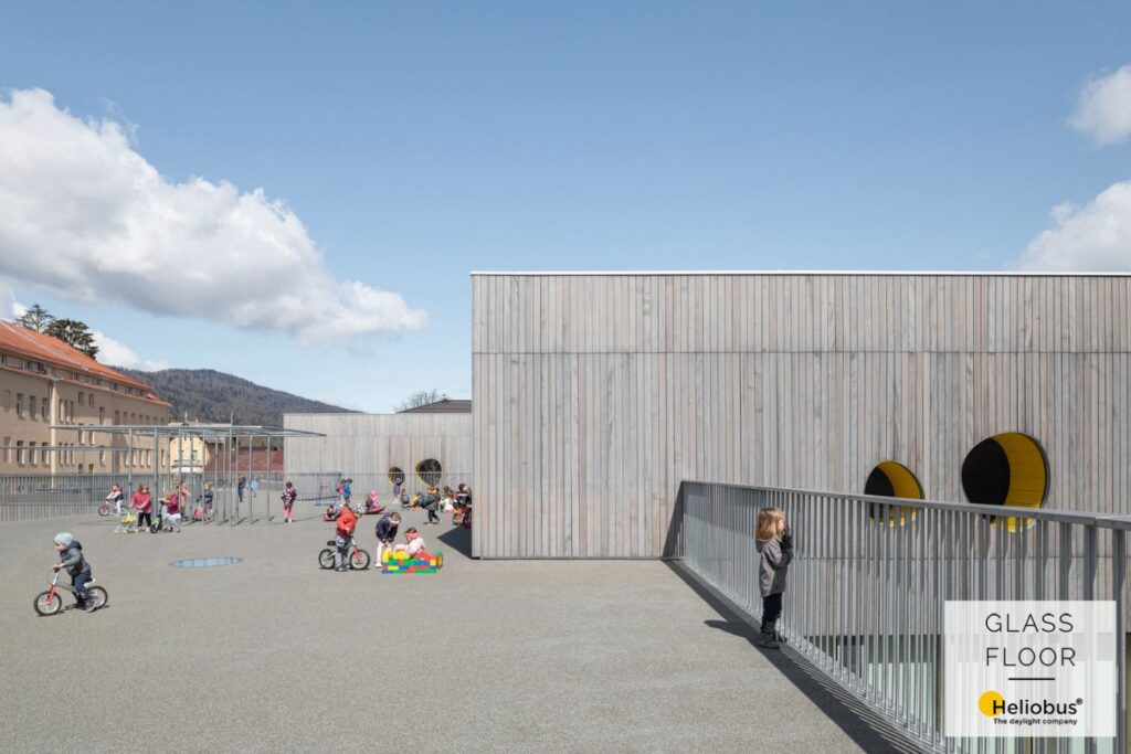 Terrasse avec aire de jeux pour enfants, où de nombreux enfants s'ébattent à vélo, tandis qu'un enfant regarde en bas, par-dessus la balustrade, dans la cour de récréation.