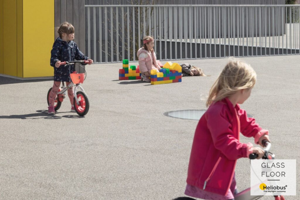 Cour de récréation d'une école maternelle avec trois enfants qui roulent à vélo et jouent avec des blocs de construction.